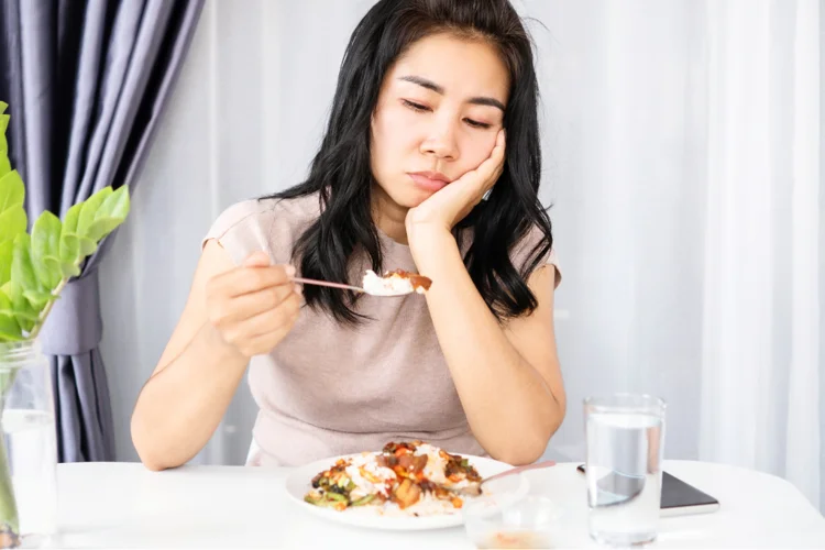 Asian woman experiencing difficulty and discomfort while chewing her food due to teeth misalignment and an overbite.