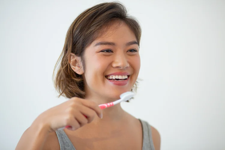 Asian woman resuming her routine of brushing teeth twice a day using a soft-bristle toothbrush and non-abrasive toothpaste after teeth whitening to maintain oral hygiene.